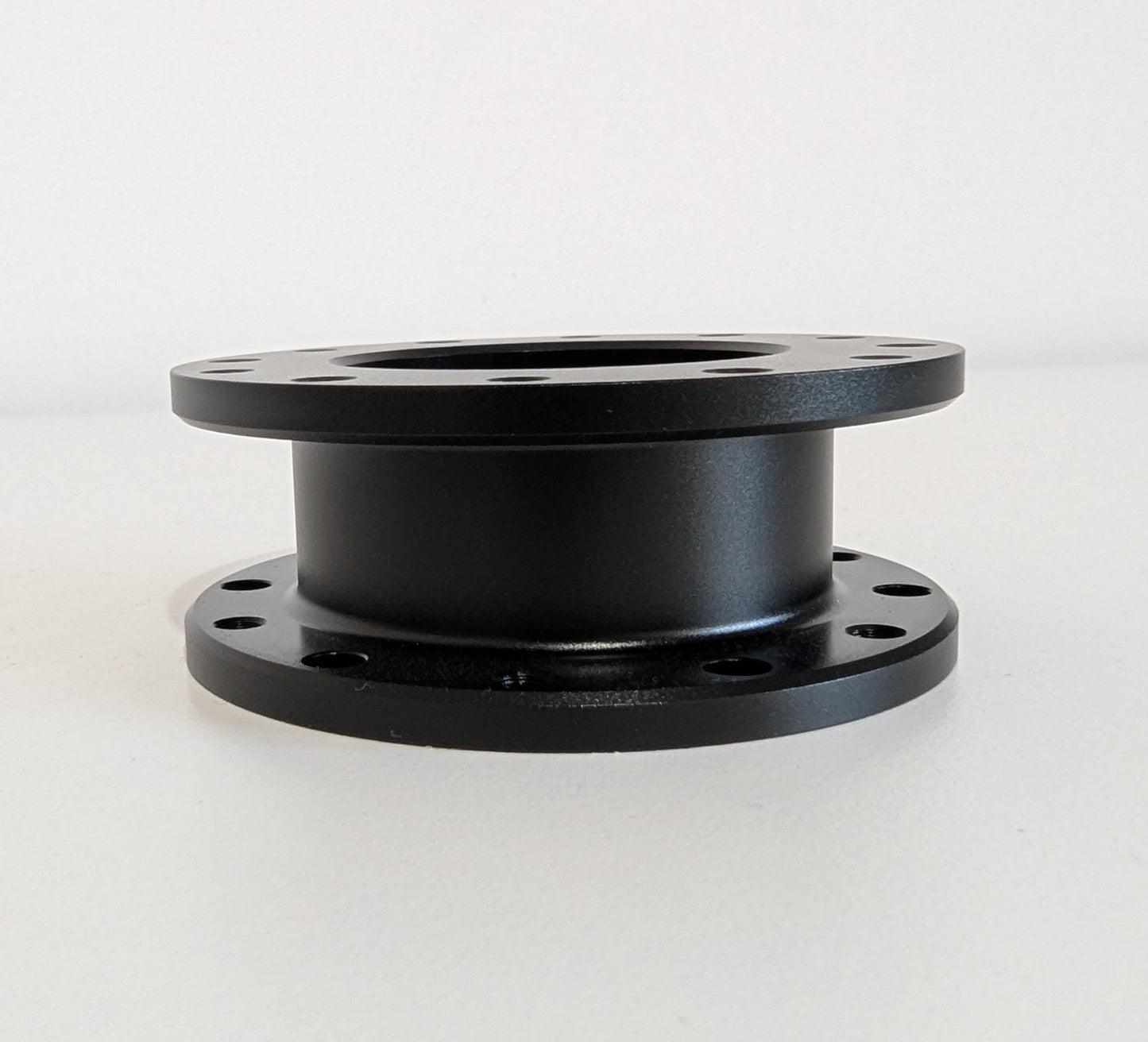 The side view of the JC 30mm Spacer. A black aluminium cylindrical object with a flange top and bottom. Bolt holes for mounting are shown around the visible diameter. Object is placed on a white surface with a white background.