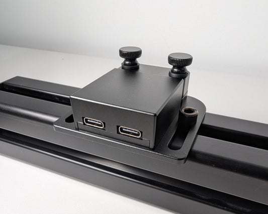 Close-up of the JC USB Power Injector mounted to black 4040 profile. The USB power injector is a black box attached to the profile with 2 bolts. The rear view shows the two black thumbscrews at the back of the image. Towards the camera you can see the two USB connections, one for power and one for PC.