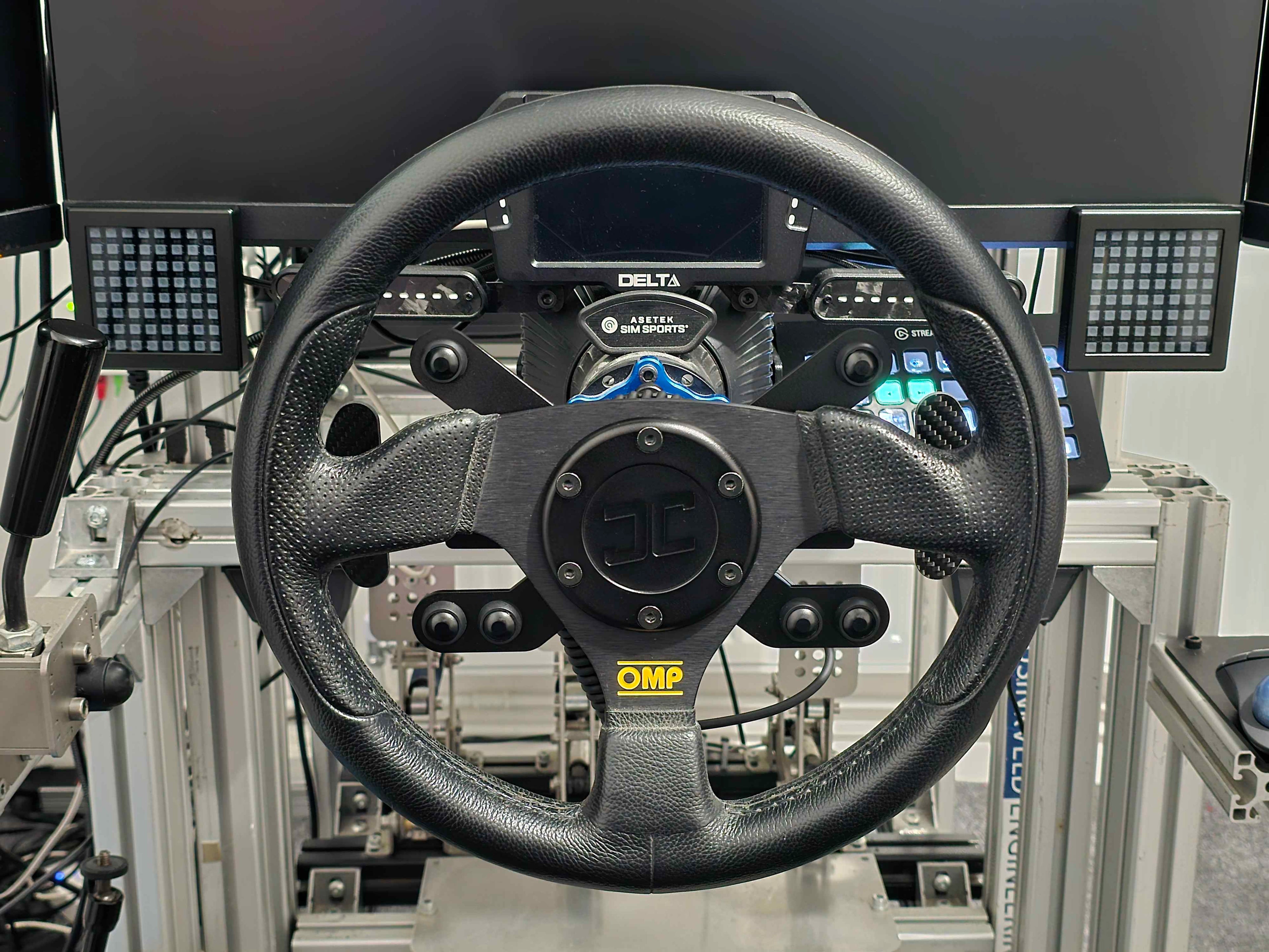 Abdul's Sim Rig setup.  A black steering wheel with the JC Wheel Hub, 6 buttons and gear shifters.  The photo also shows a monitor and two JC Sim Flags in the background.