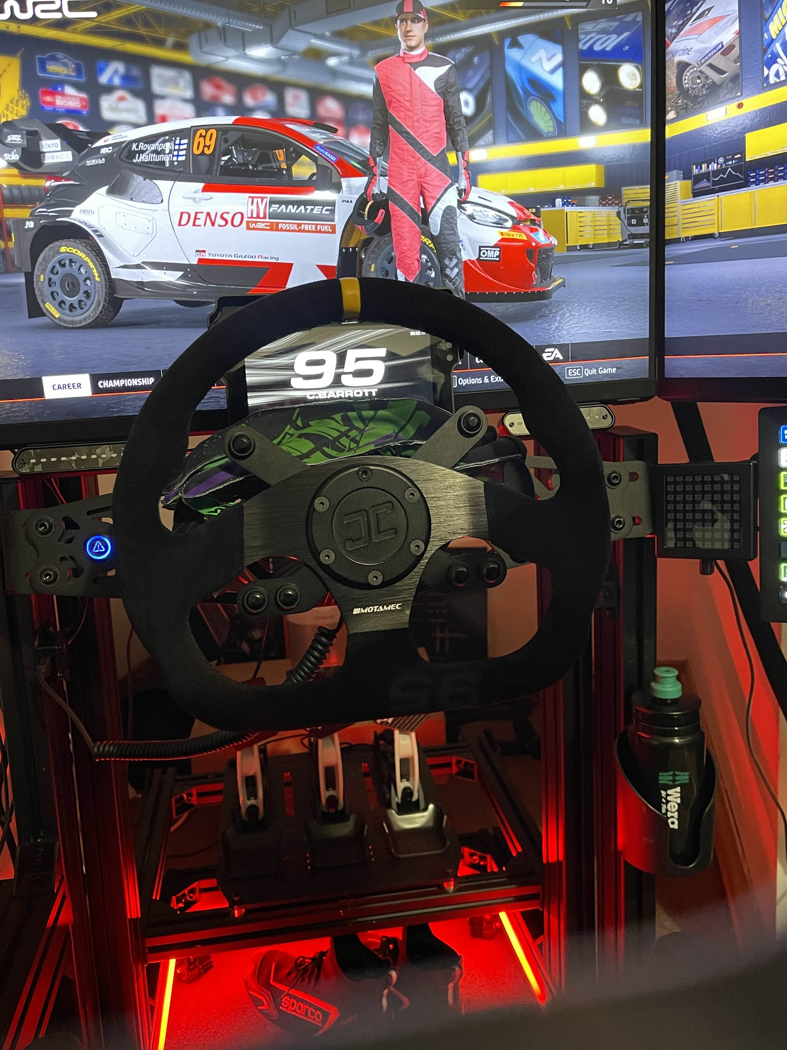 Racing simulator setup photo from Connor with steering wheel and screen displaying a racing game. The image shows the JCSS Wheel Hub, Button Brackets, Sim Flag and Power Injector.  Red lighting is used and a D Shaped Wheel.
