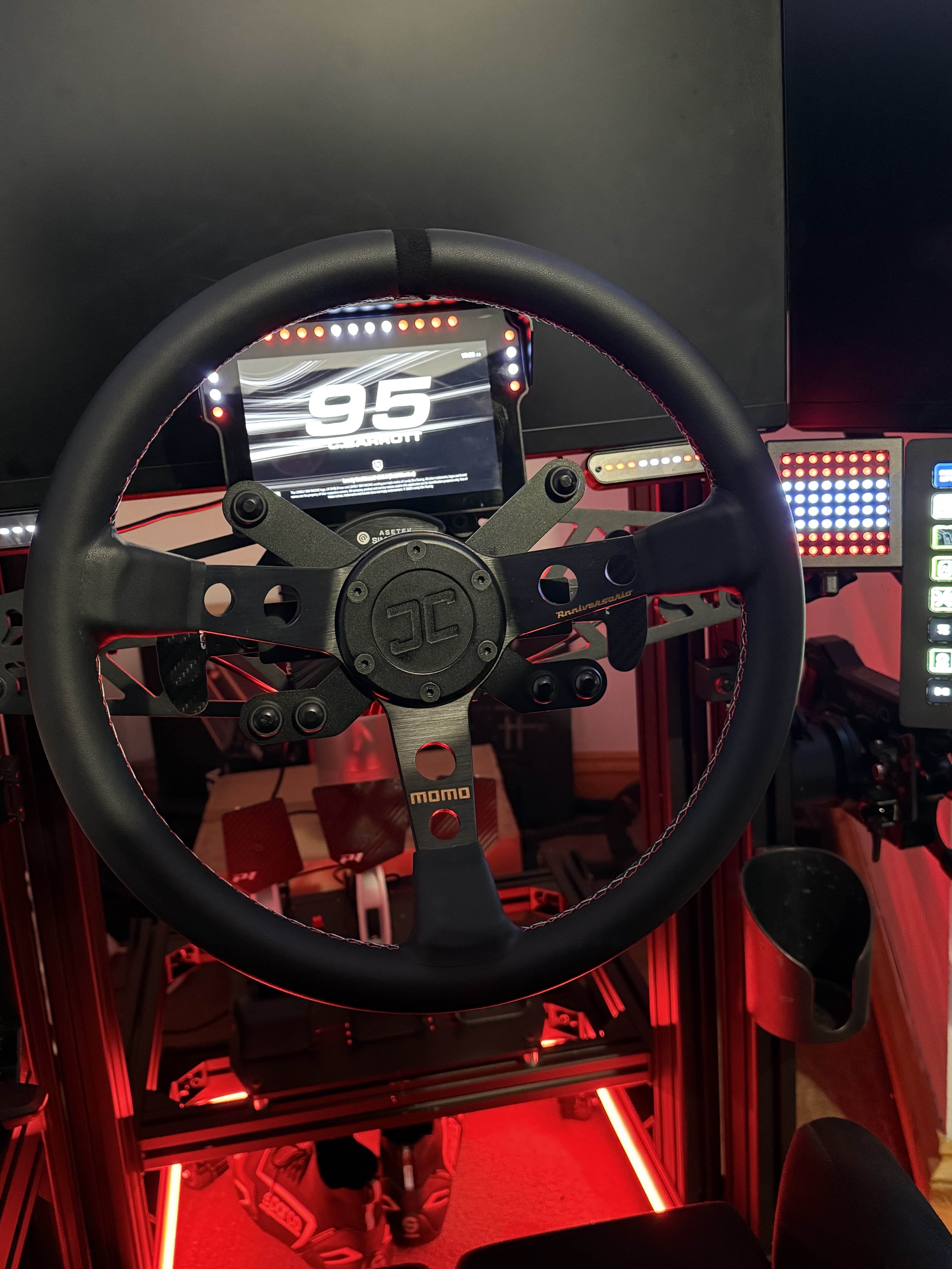 Racing simulator setup photo from Connor with steering wheel and screen displaying a racing game. The image shows the JCSS Wheel Hub, Button Brackets, Sim Flag and Power Injector.  Red lighting is used and a Deep Dish Wheel.