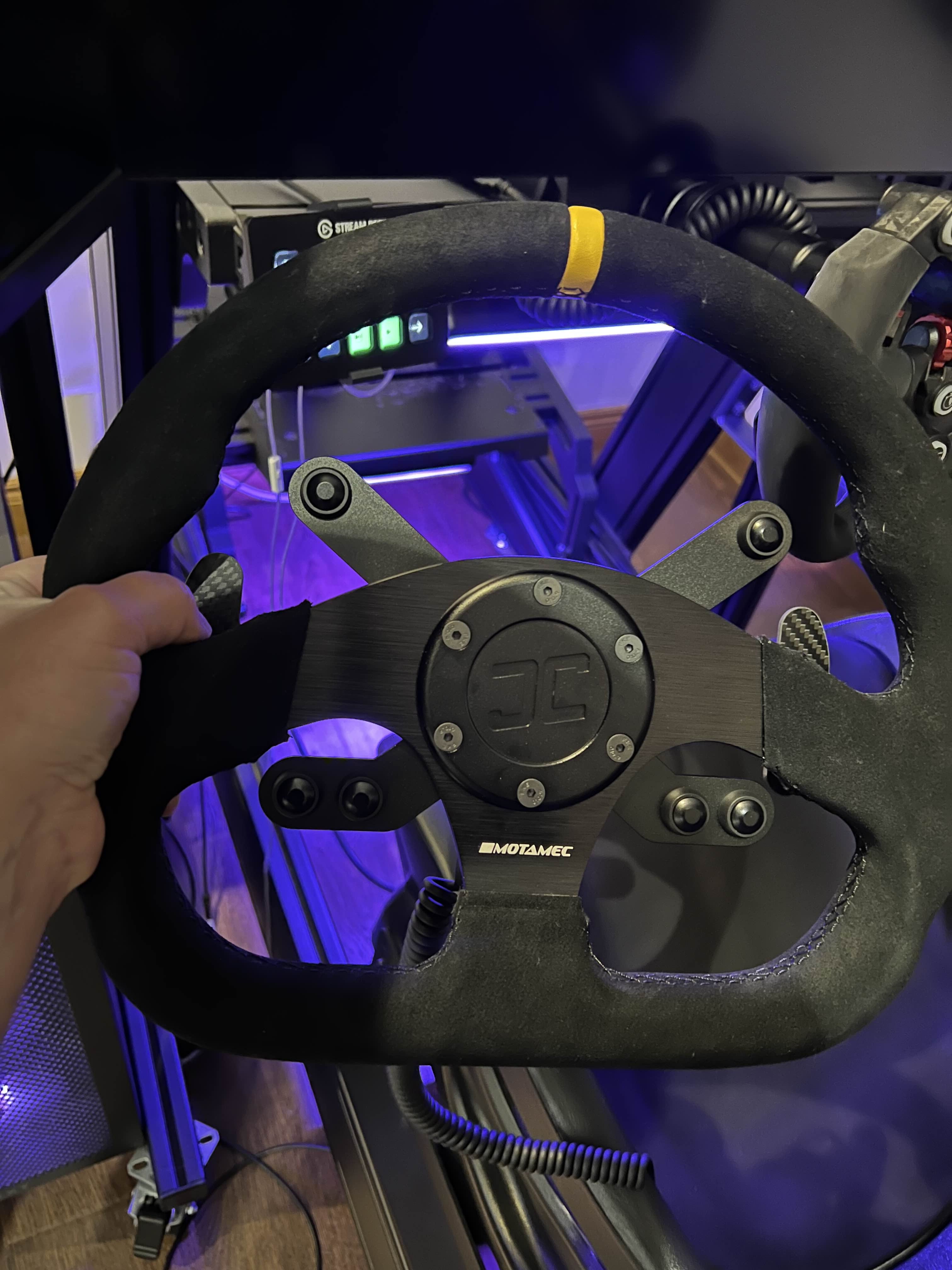 Black steering wheel with a logo held by a hand in front of a gaming setup with purple lighting.  The wheel has the JCSS Wheel hub, button brackets with 6 buttons and gear shifters attached.