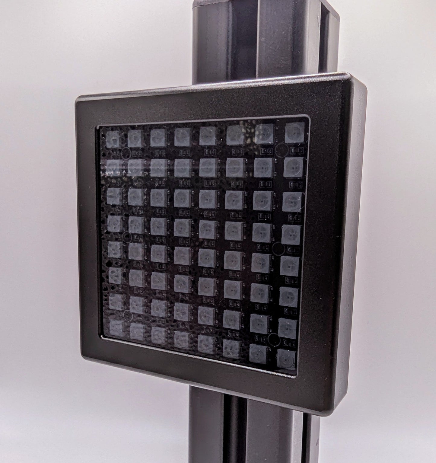 A photograph of the front of the JCSS Sim Flag mounted on a piece of black aluminium profile.  The Sim Flag is a black aluminium square with a small border, a LED light board inside under grey frosted perspex.  The LEDs are laid out in an 8x8 square.  The photograph is taken at a slight angle.