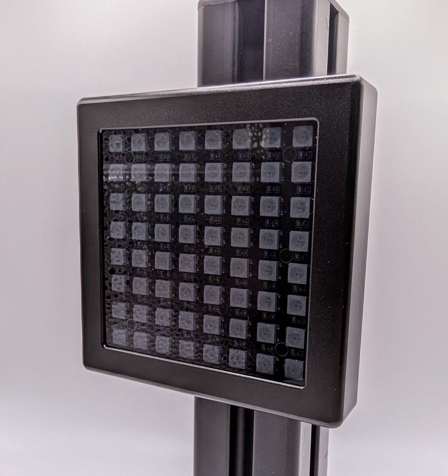 A photograph of the front of the JCSS Sim Flag mounted on a piece of black aluminium profile.  The Sim Flag is a black aluminium square with a small border, a LED light board inside under grey frosted perspex.  The LEDs are laid out in an 8x8 square.  The photograph is taken at a slight angle.