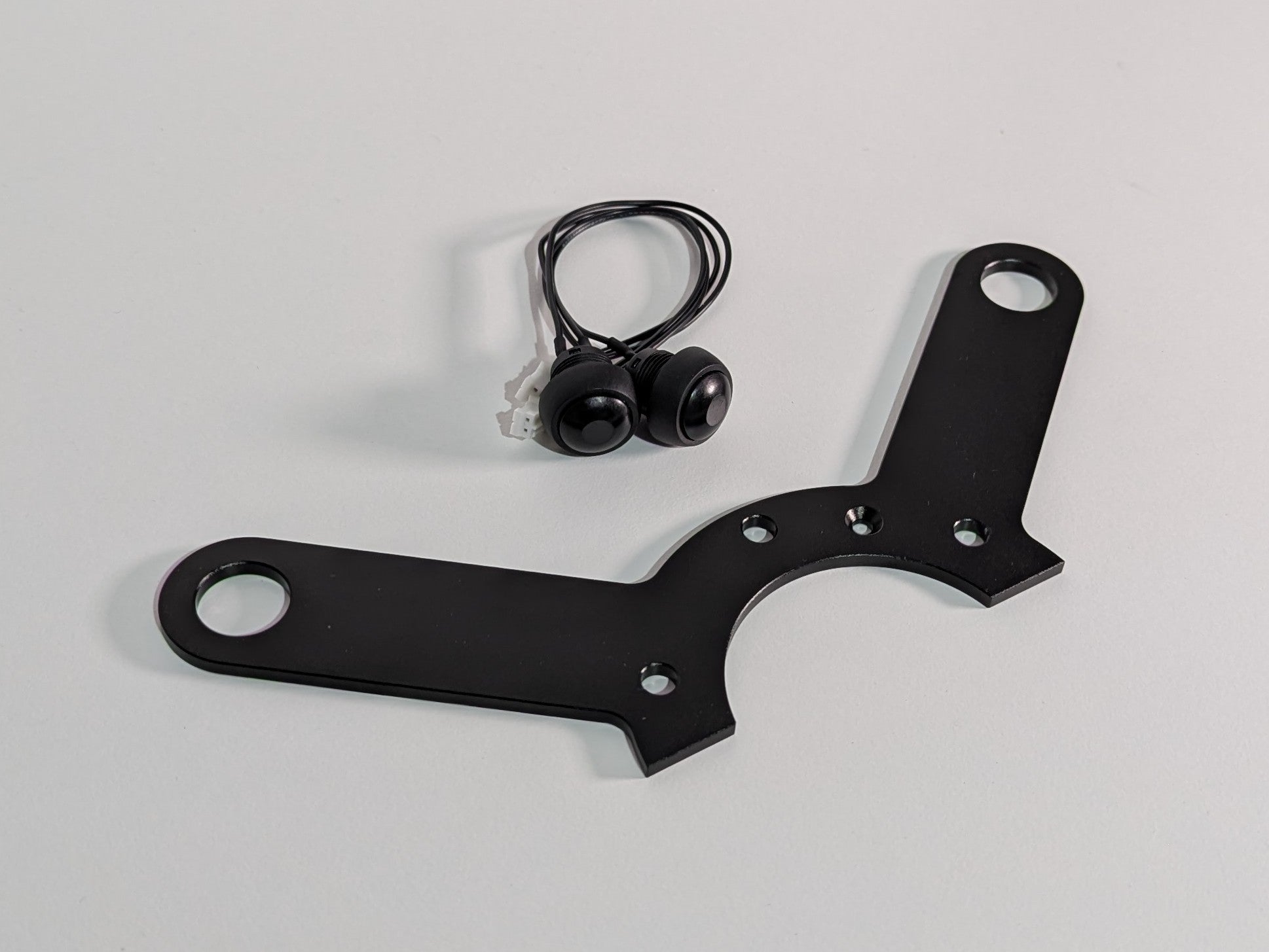 The JCSS 2 Button bracket on a white surface with two buttons and cables. The bracket is black aluminium finish with a half circular feature on the centre with two long arms with holes at each end for mounting buttons.