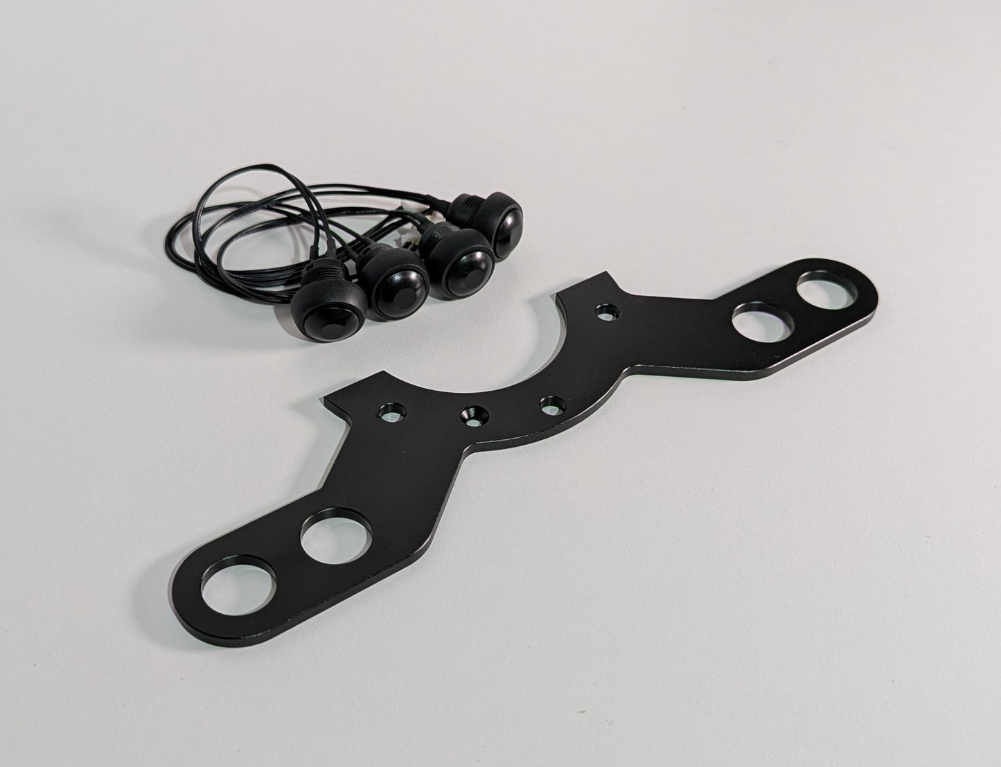 The JCSS 4 Button bracket on a white surface with four buttons and cables. The bracket is black aluminium finish with a half circular feature on the centre with two long arms with two holes at each end for mounting buttons.