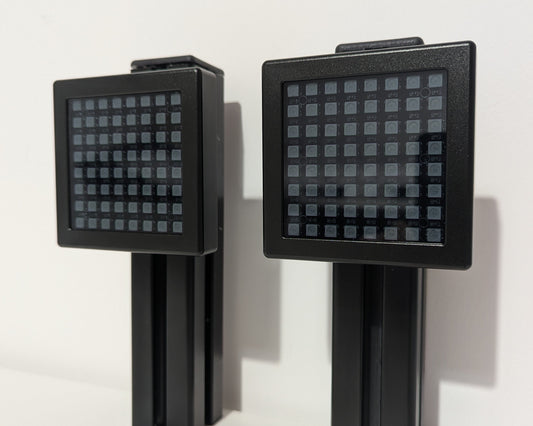 A photograph of the front of the JCSS Sim Flag mounted on a piece of black aluminium profile.  The Sim Flag is a black aluminium square with a small border, a LED light board inside under grey frosted perspex.  The LEDs are laid out in an 8x8 square.  The photograph is taken at a slight angle.