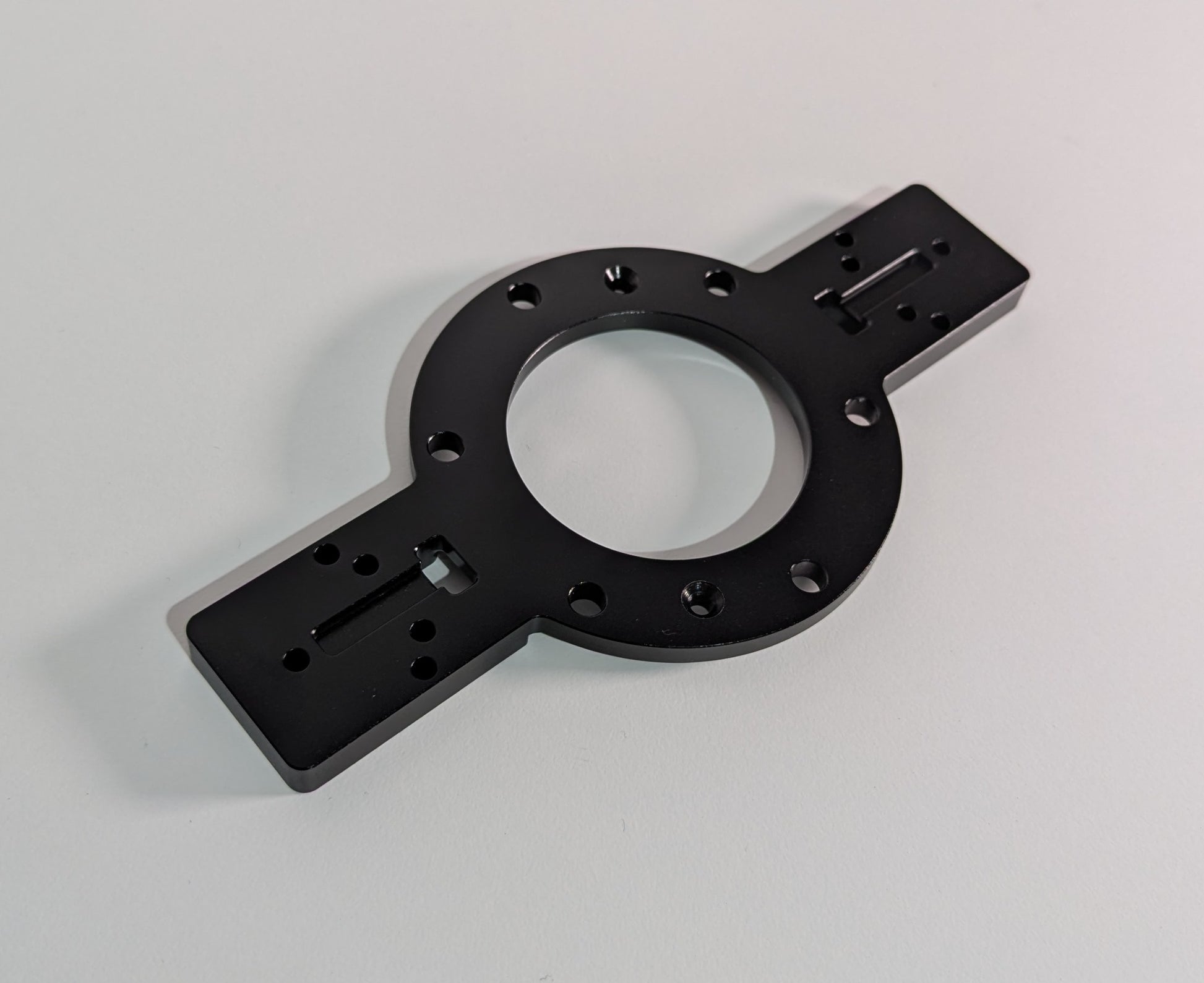 The JCSS Shifter Bracket on a white surface. The item is black anodised aluminium and comprises of a circular feature in the centre with two rectangular arms. The arms have mounting holes for shifters. The circular feature allows mounting to the JC Wheel Hub.
