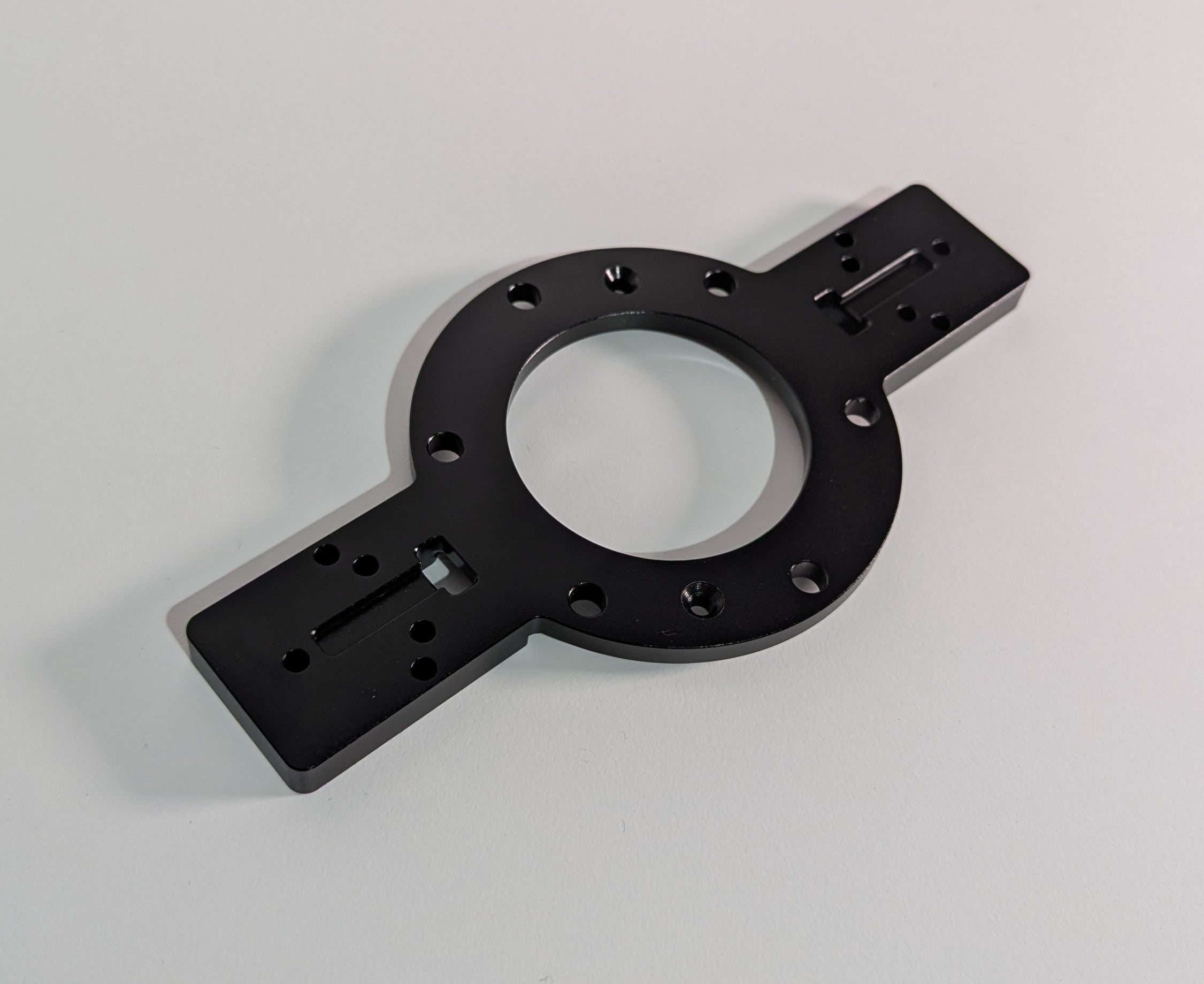 The JCSS Shifter Bracket on a white surface. The item is black anodised aluminium and comprises of a circular feature in the centre with two rectangular arms. The arms have mounting holes for shifters. The circular feature allows mounting to the JC Wheel Hub.