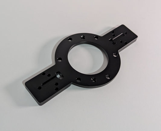 The JCSS Shifter Bracket on a white surface. The item is black anodised aluminium and comprises of a circular feature in the centre with two rectangular arms. The arms have mounting holes for shifters. The circular feature allows mounting to the JC Wheel Hub.