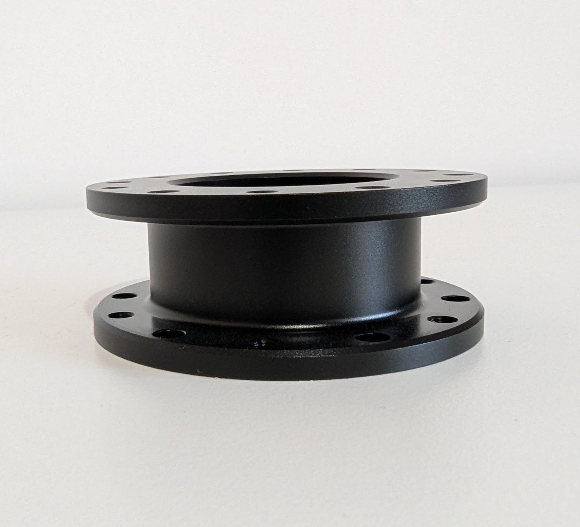 The side view of the JC 30mm Spacer.  A black aluminium cylindrical object with a flange top and bottom. Bolt holes for mounting are shown around the visible diameter.  Object is placed on a white surface with a white background.