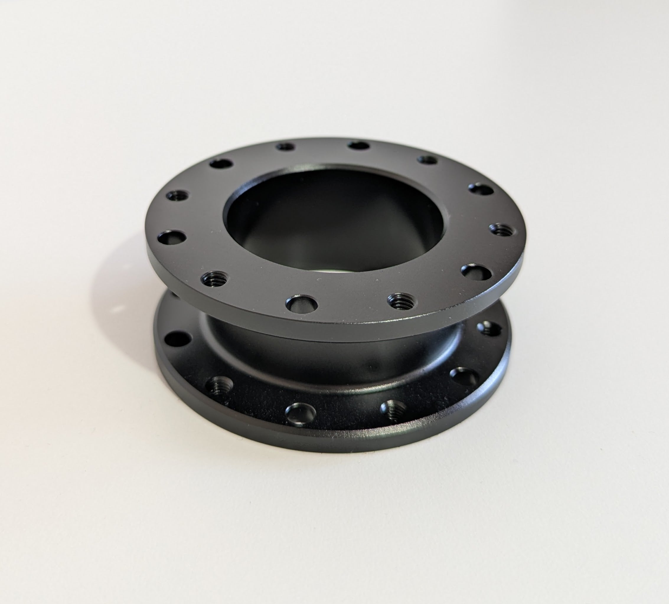 The top-down view of the JC 30mm Spacer.  A black aluminium cylindrical object with a flange top and bottom. Bolt holes for mounting are shown around the visible diameter.  Object is placed on a white surface with a white background.