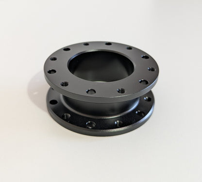 The top-down view of the JC 30mm Spacer.  A black aluminium cylindrical object with a flange top and bottom. Bolt holes for mounting are shown around the visible diameter.  Object is placed on a white surface with a white background.