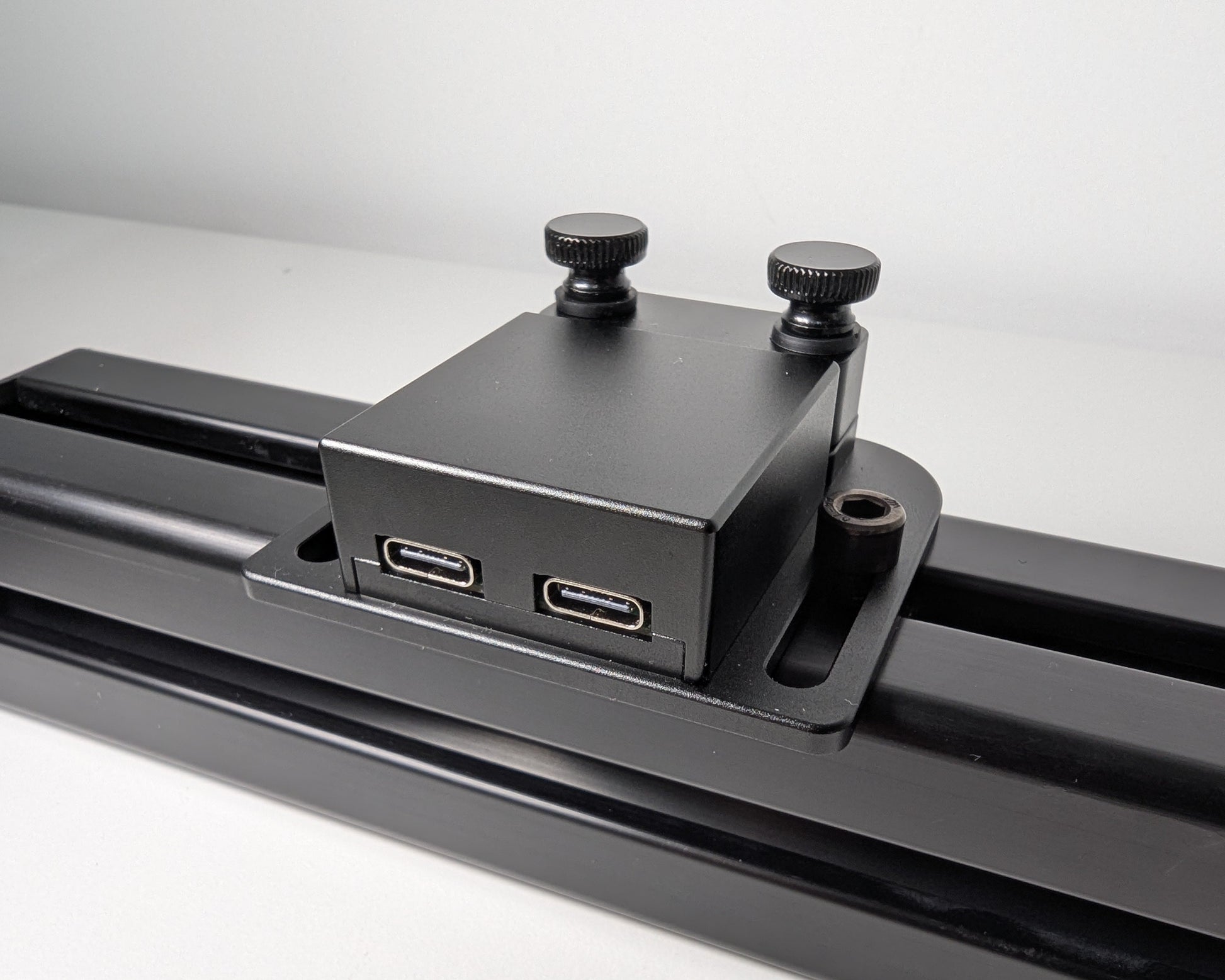 Close-up of the JC USB Power Injector mounted to black 4040 profile.  The USB power injector is a black box attached to the profile with 2 bolts.  The rear view shows the two black thumbscrews at the back of the image.  Towards the camera you can see the two USB connections, one for power and one for PC. 