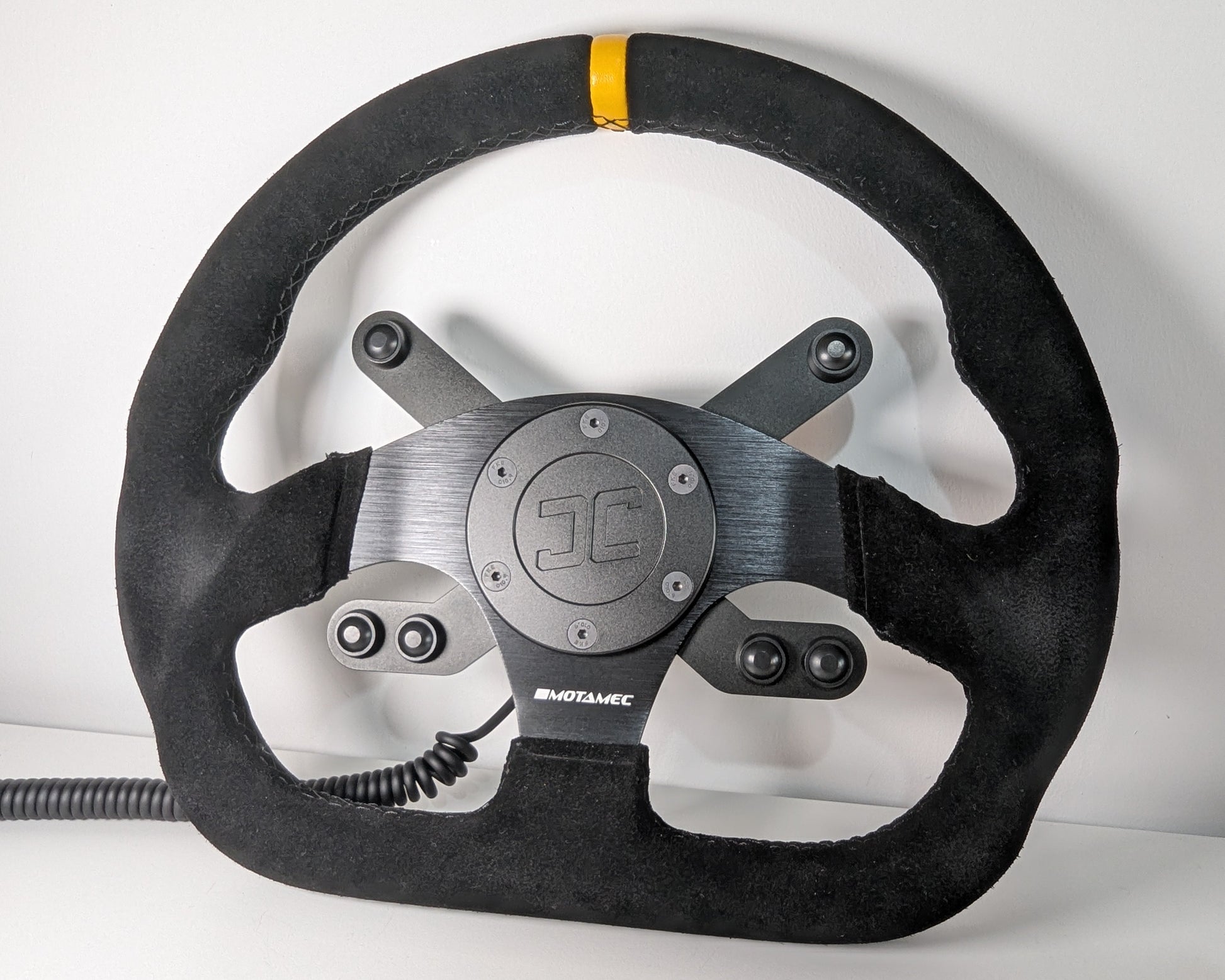 Black steering wheel with 'JC' branding on a white background  The wheel has a JC wheel hub with a top 2 button bracket and a bottom 4 button bracket with black buttons mounted.  The hub has a black coiled cable.