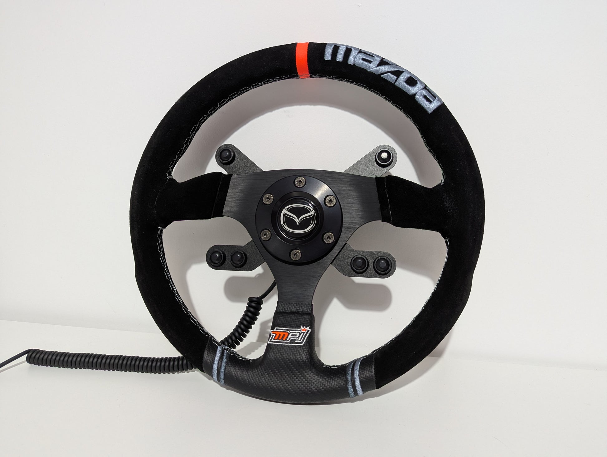 Black MPI Mazda steering wheel with 'JC' branding on a white background  The wheel has a JC wheel hub with a top 2 button bracket and a bottom 4 button bracket with black buttons mounted.  The hub has a black coiled cable.
