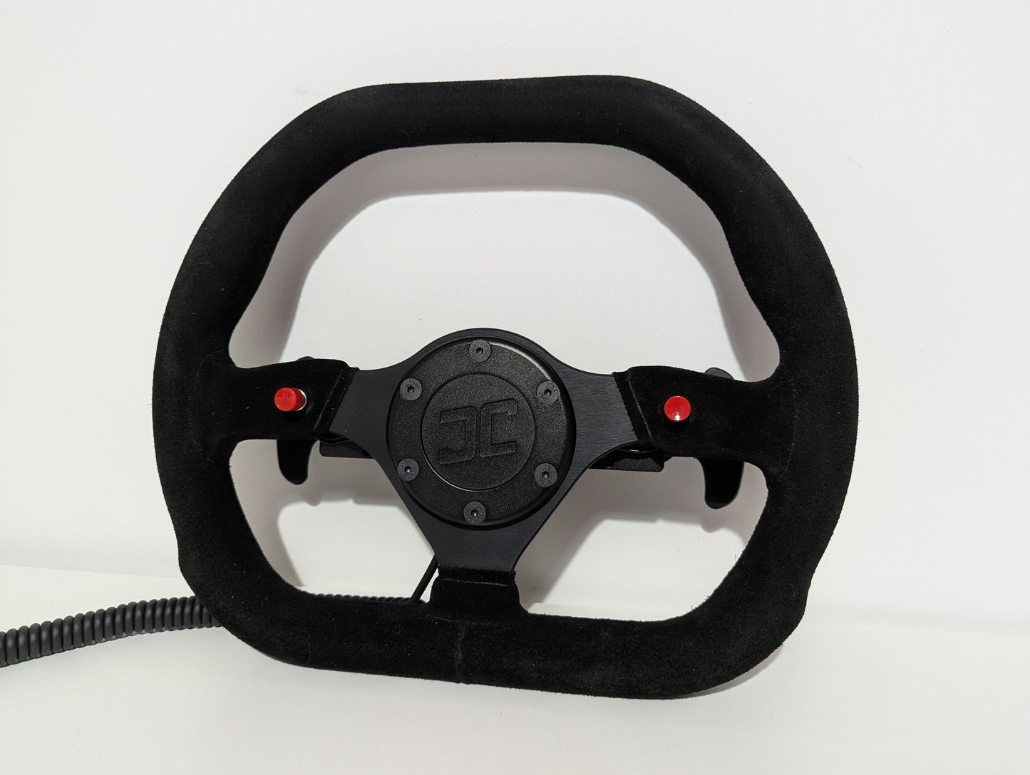 Black Sparco steering wheel with 'JC' branding on a white background  The wheel has a JC wheel hub with 2 red buttons and a set of shifters mounted on a shifter bracket.  The hub has a black coiled cable.
