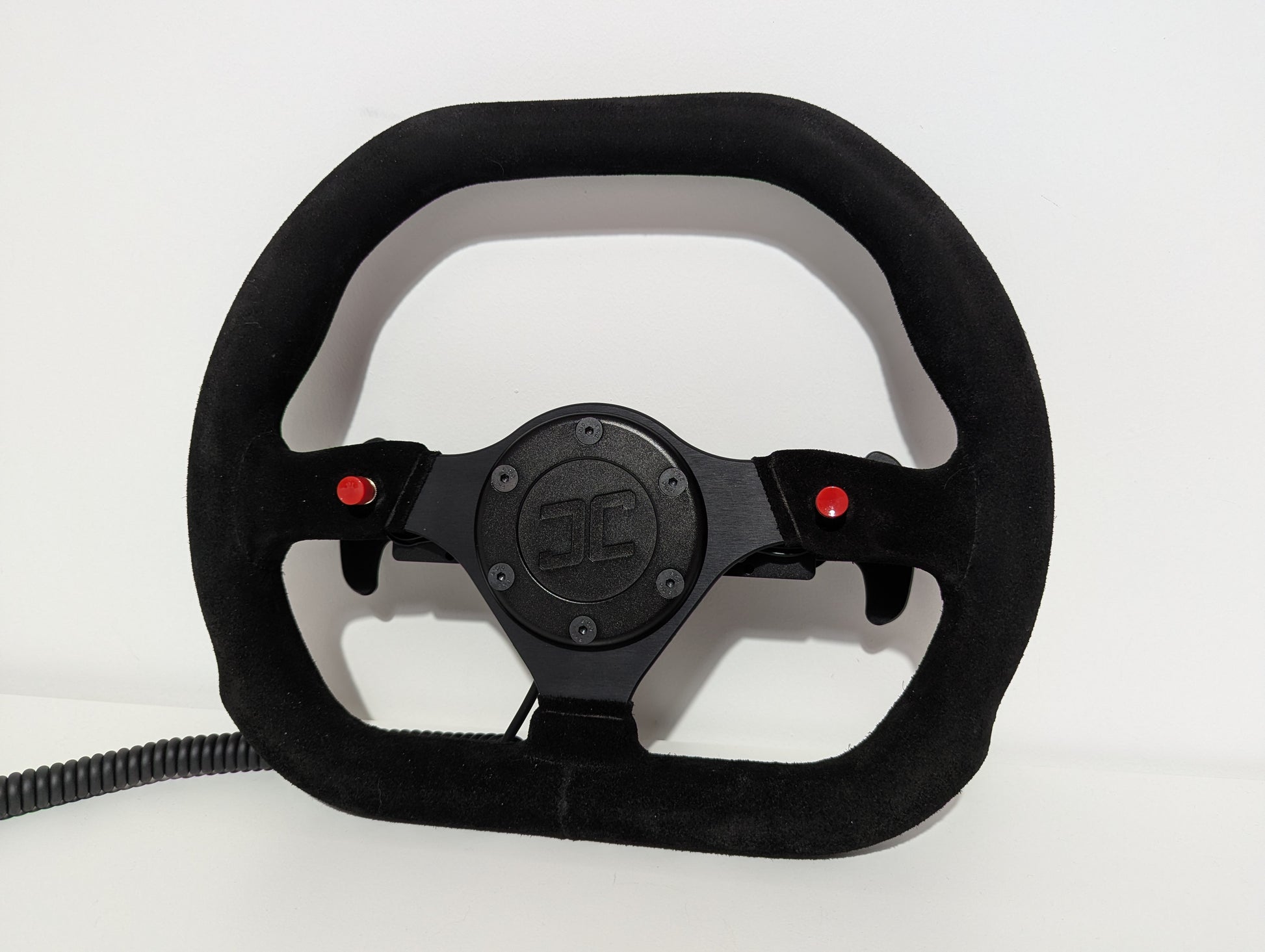 Black Sparco steering wheel with 'JC' branding on a white background  The wheel has a JC wheel hub with 2 red buttons and a set of shifters mounted on a shifter bracket.  The hub has a black coiled cable.
