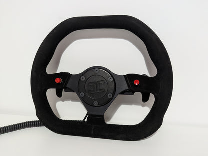 Black Sparco steering wheel with 'JC' branding on a white background  The wheel has a JC wheel hub with 2 red buttons and a set of shifters mounted on a shifter bracket.  The hub has a black coiled cable.