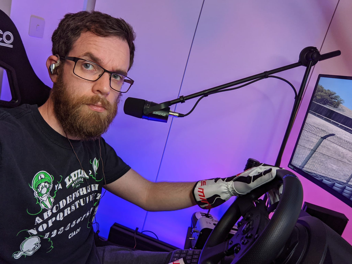 JC in a gaming rig setup with a microphone and steering wheel, wearing a black t-shirt with graphics.